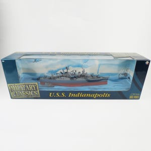 May include: A rectangular cardboard display box containing a 1:700 scale die-cast model of the U.S.S. Indianapolis warship. The box features blue and gold text, a clear plastic window, and a printed maritime scene behind the gray and red ship.