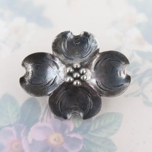 May include: A silver-toned metal brooch in the shape of a dogwood flower. The brooch features four petals with curved edges and a cluster of small, round accents at the center. The metal has a slightly aged appearance.