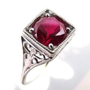 May include: A silver ring featuring a large, square-cut, deep red gemstone. The setting has a decorative, filigree design on the sides and small, round accents around the gemstone. The ring is set against a white background.