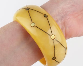 Könnte beinhalten: Ein breites, gelb getöntes Armreif-Armband mit geometrischem Design. Das Armband hat eine Reihe kleiner, goldfarbener Kreise, die durch dünne, dunkle Linien verbunden sind. Das Armband wird am Handgelenk getragen.