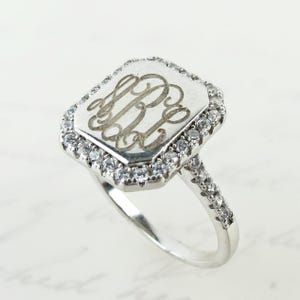 May include: A silver ring with a rectangular face featuring an engraved monogram. The face is surrounded by a halo of small, clear gemstones. The ring's band is also adorned with similar gemstones, creating a sparkling effect.