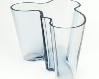 May include: A clear, light blue glass vase featuring an organic, undulating shape with multiple curved sections. The vessel has a smooth, transparent surface and a wide, open top, showcasing a fluid, asymmetrical silhouette from a high angle.