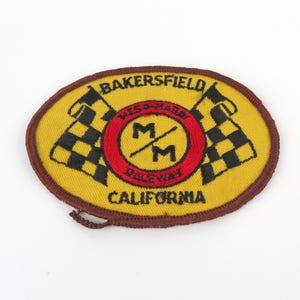 May include: Oval-shaped patch with a yellow background, featuring black and white checkered flags on either side of a red circle with the letters "MM". The patch reads "BAKERSFIELD RACEWAY CALIFORNIA".