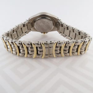 May include: A silver-toned wristwatch with a two-tone band. The watch band features silver links with gold-colored accents. The watch face is round and silver, with a silver and gold-toned band. The watch is displayed on a white surface.