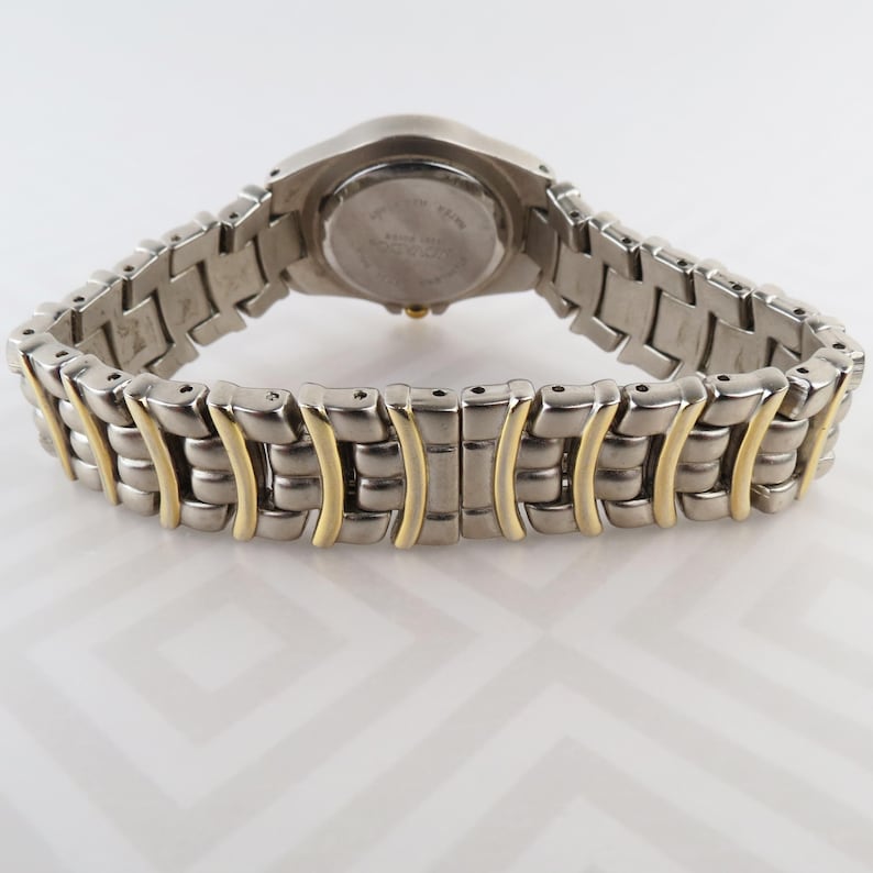 May include: A silver-toned wristwatch with a two-tone band. The watch band features silver links with gold-colored accents. The watch face is round and silver, with a silver and gold-toned band. The watch is displayed on a white surface.