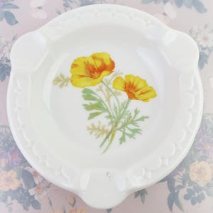 May include: White ceramic ashtray with a floral design. The ashtray features a detailed illustration of yellow and orange poppies with green leaves. The rim has a scalloped edge and three resting points.