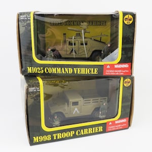 May include: Two toy military vehicles in boxes. The top box reads "M1025 COMMAND VEHICLE" and the bottom box reads "M998 TROOP CARRIER". Each box contains a miniature vehicle and a soldier figurine. The boxes have a camouflage design.