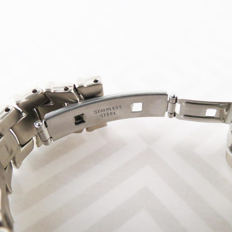 May include: Close-up of a stainless steel watch band clasp. The clasp is open, revealing the words "STAINLESS STEEL" engraved on the metal. The band is made of interlocking metal links.