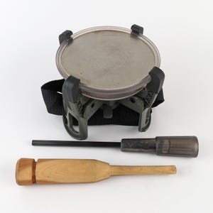May include: A camping stove with a clear, round top and a black strap. The stove has a dark green base with black clips. A black rod and a wooden tool are also included.