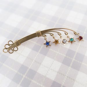 May include: A gold-toned brooch with a decorative design. The brooch features a series of curved lines with dangling star-shaped charms, each holding a different colored gemstone: blue, yellow, clear, green, and red. The brooch is set against a plaid background.