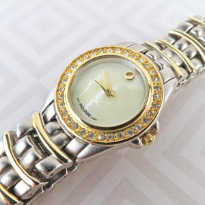 May include: A Movado quartz watch with a silver and gold-tone stainless steel band. The watch face is a light cream color, surrounded by a gold bezel set with small, clear crystals. The watch hands are gold.