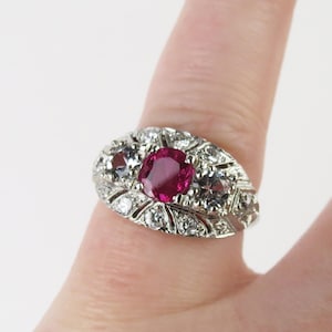 May include: A vintage-style ring featuring a central oval ruby gemstone, surrounded by smaller clear gemstones. The ring has an ornate, detailed design with a silver or white gold band.