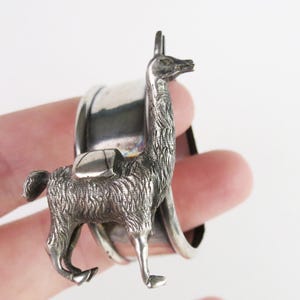 May include: A silver-toned llama napkin ring. The llama has detailed fur and a saddle. The ring is a curved, polished metal. The llama stands upright, with its head held high. The design is intricate and detailed, suitable for table decor.