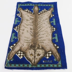 May include: A rectangular fabric patch featuring a central illustration of a grey wolf pelt on a vibrant blue background. The design includes a black and white checkered border, teal geometric accents, and the word OLLAN printed in small letters.