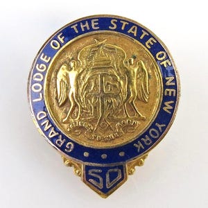 May include: A gold-toned pin with blue enamel trim. The pin features the words "GRAND LODGE OF THE STATE OF NEW YORK" and a central emblem with two figures and a shield. A small blue banner at the bottom displays the number "50".