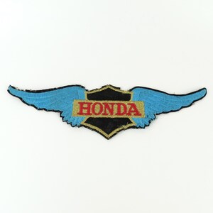 May include: Embroidered patch featuring the Honda logo with a winged design. The patch has a black and gold shield shape with the word "HONDA" in red. The wings are blue with black outlining.