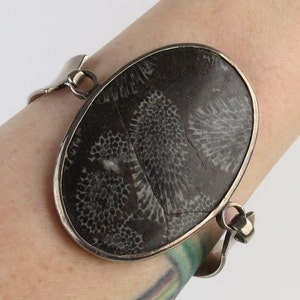 May include: A silver cuff bracelet featuring a large, oval-shaped stone with a dark gray and white coral pattern. The stone is set in a silver bezel and the bracelet has a curved, open design.