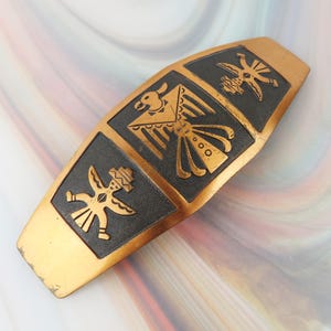 May include: A gold-toned metal hair barrette featuring a central rectangular panel with a black textured background. The panel displays three engraved figures, including a large central bird-like motif flanked by two smaller stylized human-like forms.