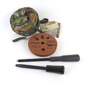 May include: A turkey hunting kit featuring a camouflage carrying case, a wooden turkey call with the text "Turkey Tracer", and two black tools. The wooden call has the text "Lohman Buck Harris" and "Yelp, Purr, Putt, Tune".