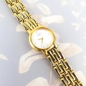 May include: A gold-tone Seiko Quartz wristwatch featuring a round white dial with gold-tone hands and hour markers. The watch has a polished gold-tone metal link bracelet with a repeating rectangular pattern and a secure clasp mechanism.
