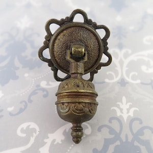 May include: Antique brass-toned drawer pull with a decorative design. The pull features a round backplate with an ornate, star-like frame. The handle has a detailed, bulbous shape with a decorative finial. A vintage-style hardware element.
