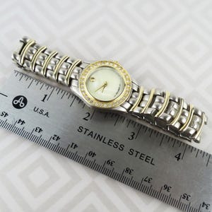 May include: A silver and gold-toned wristwatch with a round, cream-colored face. The watch has a gold bezel encrusted with small, clear crystals. The band is silver with gold accents. The watch is next to a stainless steel ruler.