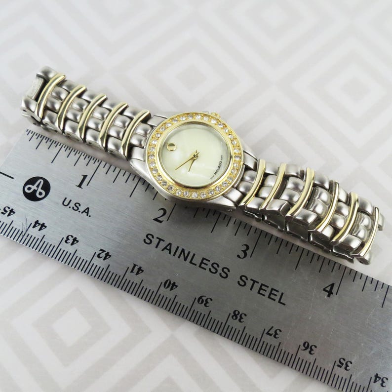 May include: A silver and gold-toned wristwatch with a round, cream-colored face. The watch has a gold bezel encrusted with small, clear crystals. The band is silver with gold accents. The watch is next to a stainless steel ruler.