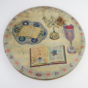 May include: A round, vintage Passover Seder plate with illustrations of traditional items. The plate features a Seder plate, a menorah, a chalice, and an open book. The border is decorated with Hebrew text and small figures.