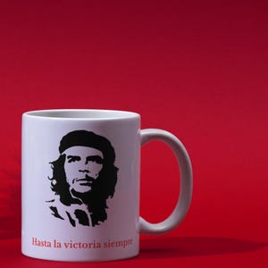May include: White ceramic mug featuring a black silhouette of a man's portrait. The mug has the text "Hasta la victoria siempre" in red. The mug is set against a solid red background.