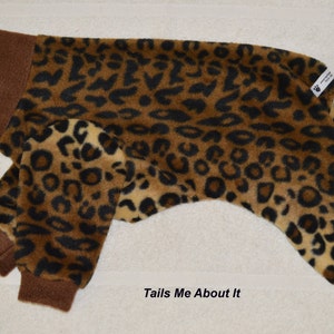 May include: Brown and tan leopard print fleece dog pajamas with brown ribbed cuffs. The pajamas have the text "Tails Me About It" printed on the bottom.
