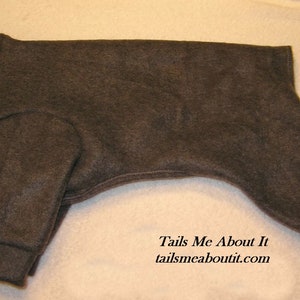 May include: A dark gray fleece dog jumpsuit with a high neck and long sleeves. The jumpsuit has a ribbed cuff at the wrists and ankles. The text "Tails Me About It" and "tailsmeaboutit.com" is printed on the bottom of the jumpsuit.