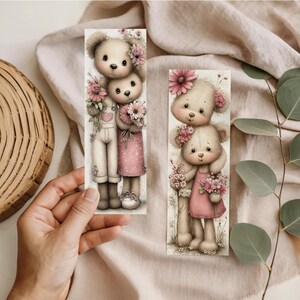 May include: Two illustrated bookmarks featuring teddy bears. One bookmark shows two bears, one in overalls and the other in a pink dress, holding flowers. The other bookmark shows two bears in a pink dress, holding flowers. The background is a light beige.