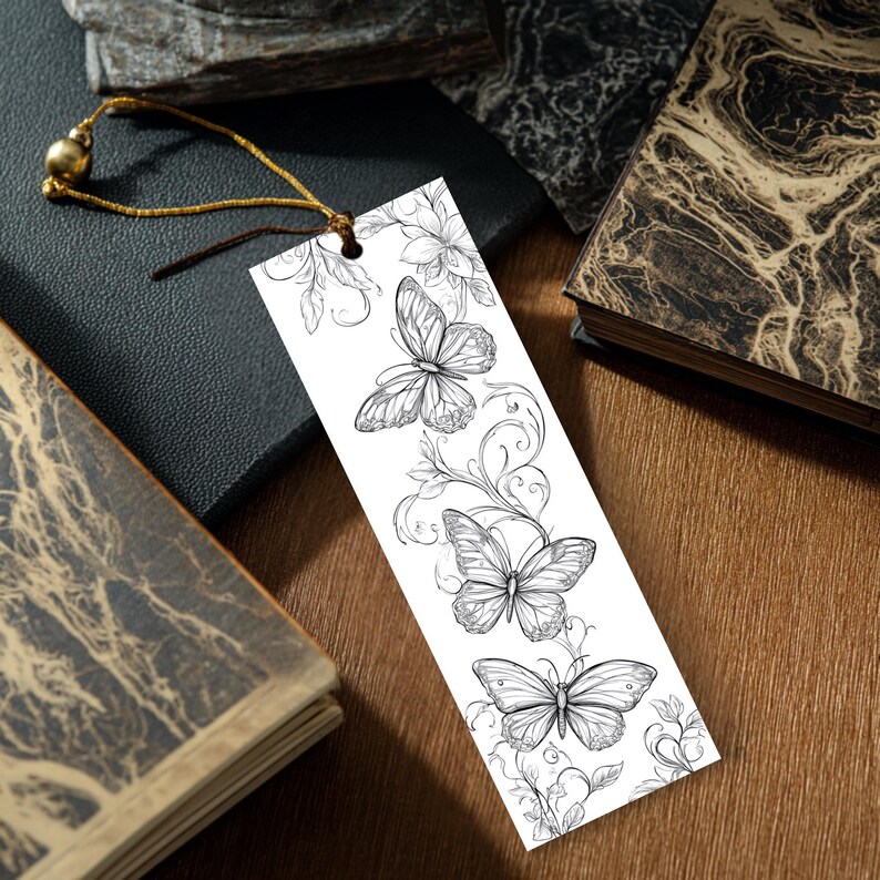 Op de afbeelding: Een rechthoekige witte boekenlegger met een zwarte inkttekening van drie vlinders en bloemranken. De boekenlegger heeft een gaatje aan de bovenkant met een dun gouden koordje, afgewerkt met een kleine, ronde, metalen gouden kraal.