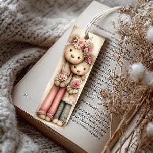 May include: A whimsical bookmark featuring two teddy bears adorned with floral crowns and holding flowers. The bears wear pink overalls and a floral dress. A white string is attached at the top. The bookmark is resting on an open book.
