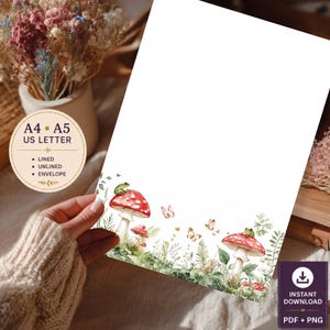 May include: A white stationery sheet featuring a watercolor illustration at the bottom with two red and white spotted mushrooms, green ferns, small plants, butterflies, and two small green frogs. Text on the circular overlay reads A4, A5, US LETTER, LINED, UNLINED, ENVELOPE.