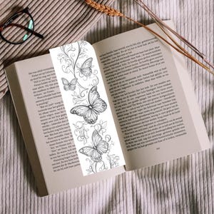Op de afbeelding: Een verticale boekenlegger met een zwart-witte lijntekening van drie vlinders omringd door wervelende bloemranken. De boekenlegger ligt in een open boek met gedrukte tekst, rustend op een oppervlak van textuurrijke stof.