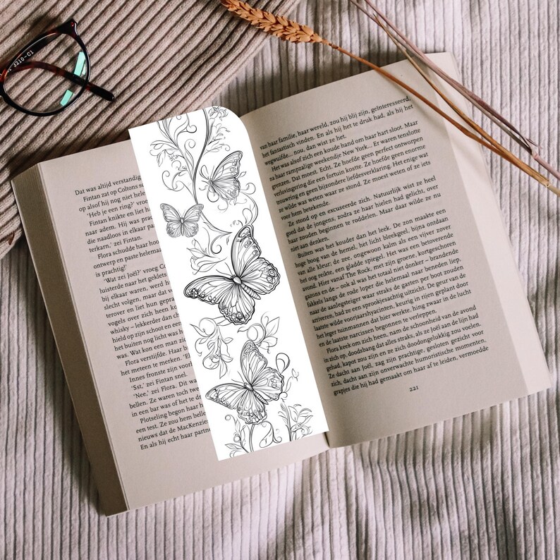 Op de afbeelding: Een verticale boekenlegger met een zwart-witte lijntekening van drie vlinders omringd door wervelende bloemranken. De boekenlegger ligt in een open boek met gedrukte tekst, rustend op een oppervlak van textuurrijke stof.