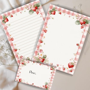 May include: Stationery set featuring a lined sheet, a blank sheet, and a card. Each piece has a white background with a pink gingham border and strawberry and floral accents. The card reads "Dear,"