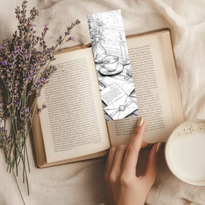 May include: A black and white bookmark featuring a cozy scene with a teacup, candle, and letters, placed in an open book. A sprig of lavender and a cup of coffee are also visible.
