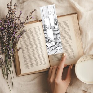 May include: A black and white bookmark featuring three candles, pinecones, and books. The bookmark is placed in an open book with a cup of coffee and lavender in the background. The bookmark design is a line drawing.