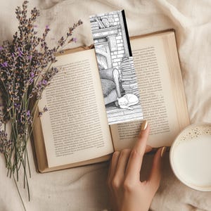 May include: A black and white bookmark featuring a cozy scene with a fireplace, armchair, and sleeping cat, inserted into an open book. A sprig of lavender and a cup of coffee with milk are also visible.
