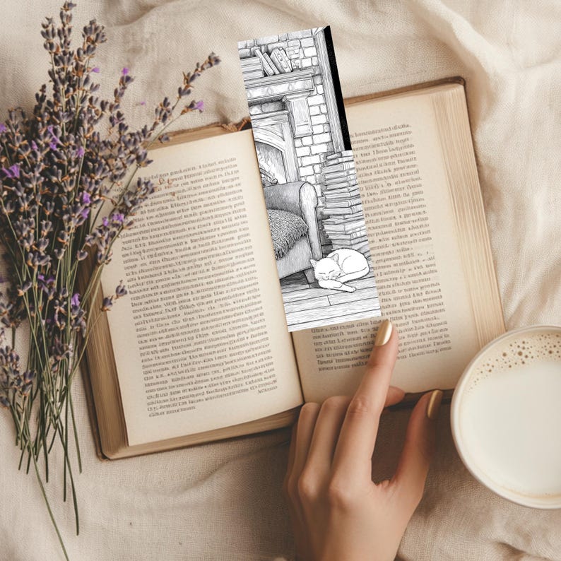 May include: A black and white bookmark featuring a cozy scene with a fireplace, armchair, and sleeping cat, inserted into an open book. A sprig of lavender and a cup of coffee with milk are also visible.