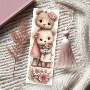 May include: A decorative bookmark featuring two teddy bears with pink flower accents. The bears are dressed in pink and white dresses and holding flowers. A pink tassel hangs from the top. The bookmark is set against a white knitted background.