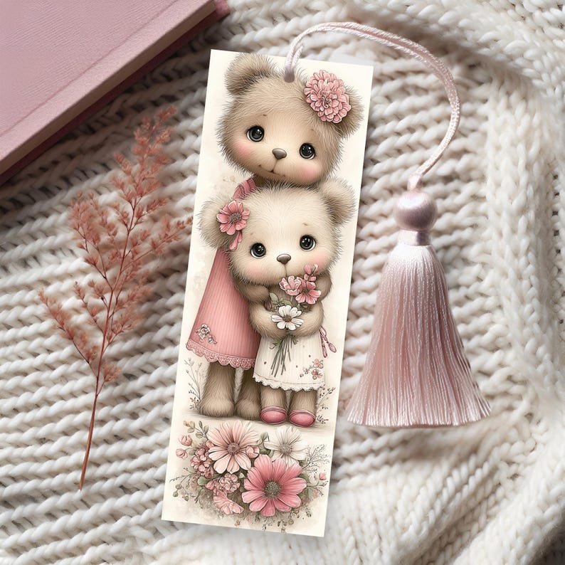 May include: A decorative bookmark featuring two teddy bears with pink flower accents. The bears are dressed in pink and white dresses and holding flowers. A pink tassel hangs from the top. The bookmark is set against a white knitted background.
