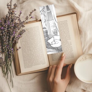 May include: A black and white illustrated bookmark depicting a cozy winter scene with a sleeping cat, fairy lights, and a snowy window. The bookmark is placed in an open book, next to a cup of coffee and a sprig of lavender.