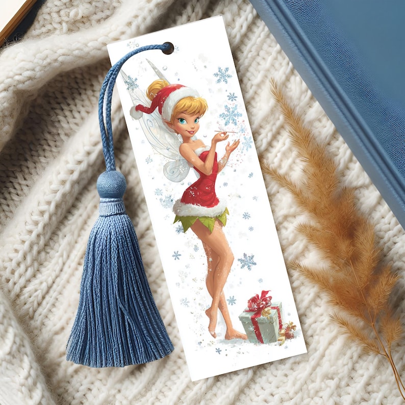 Peut inclure: Un marque-page rectangulaire en papier illustr&eacute; d'une f&eacute;e blonde v&ecirc;tue d'une tenue de style P&egrave;re No&euml;l rouge et blanche avec des bordures vertes. La f&eacute;e tient une baguette parmi des flocons bleus. Un cordon bleu avec un pompon est attach&eacute;.