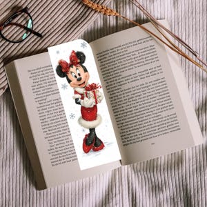 Peut inclure: Un marque-page vertical illustr&eacute; d'une souris de dessin anim&eacute; portant une robe rouge &agrave; bordure en fourrure blanche, un n&oelig;ud rouge assorti et des chaussures rouges. Elle tient un cadeau, entour&eacute;e de flocons de neige sur fond blanc.