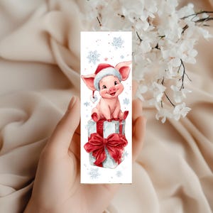 Peut inclure: Un marque-page rectangulaire pr&eacute;sentant une illustration &agrave; l'aquarelle d'un porcelet rose portant un bonnet de P&egrave;re No&euml;l rouge et blanc. Le porcelet est assis sur un cadeau blanc orn&eacute; d'un grand ruban rouge, entour&eacute; de flocons de neige bleus.
