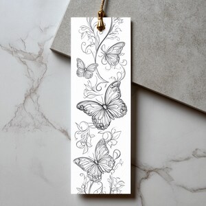 Op de afbeelding: Een rechthoekige witte boekenlegger met een zwarte lijntekening van drie vlinders omringd door zwierige bloemranken. De boekenlegger heeft aan de bovenkant een goudkleurig metalen oogje met een dun, lichtbruin koordje eraan bevestigd.