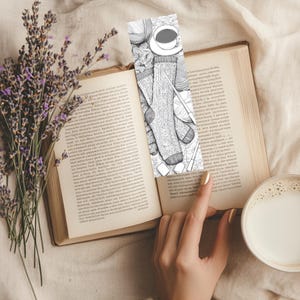 May include: A bookmark featuring a black and white illustration of knitting supplies, including socks, yarn, and a cup of coffee, rests in an open book. A sprig of lavender and a cup of coffee are also in the image.